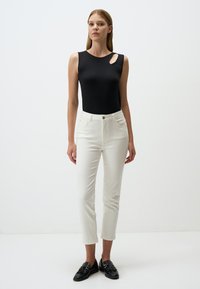 Bodycon nero senza maniche con aperture sulle spalle, abbinato a jeans bianchi slim-fit e mocassini neri, presentato su uno sfondo neutro.
