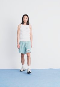 Fehér trikó és világoskék rövidnadrág Nike logóval, fehér és fekete sportcipővel párosítva, kék padlón állva, világos háttér előtt.