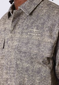 Veste chemise à motif gris et beige avec col, poches poitrine et détails de logo subtils. Tissu texturé avec un aspect robuste.