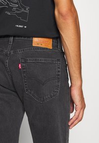 Schwarze Jeans aus Denim mit einem ausgeprägten Designelement an der Gesäßtasche, ausgestattet mit einem Lederlabel am Bund und einem roten Flicken an der Tasche.