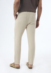 Beige, leichte Hose aus strukturiertem Stoff, Slim Fit, mit zwei Gesäßtaschen. Kombiniert mit lässigen Sandalen, von hinten gezeigt.