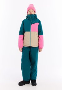 Barevný lyžařský outfit skládající se z tyrkysové, růžové a béžové bundy s vysokým límcem a zipem, doplněný o odpovídající tyrkysové kalhoty a černé boty.