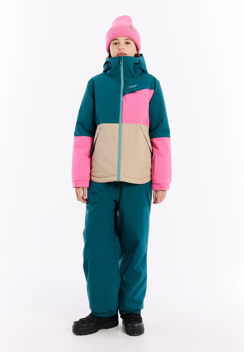 Barevný lyžařský outfit skládající se z tyrkysové, růžové a béžové bundy s vysokým límcem a zipem, doplněný o odpovídající tyrkysové kalhoty a černé boty.