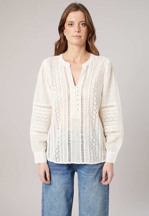 Vrouw draagt een witte blouse met lange mouwen, kanten patronen en knopen aan de voorkant, gecombineerd met een blauwe jeans tegen een effen achtergrond.