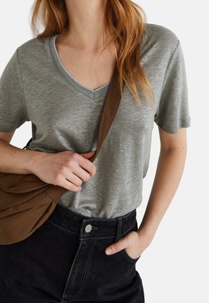 T-shirt gris à col en V avec un léger éclat, associé à un jean noir taille haute. Un sac à bandoulière marron repose contre le corps.