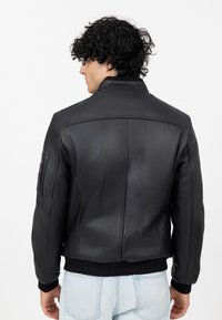 Giacca di pelle nera con collare alto, polsini a coste e una tasca con zip sulla manica sinistra; texture liscia e design aderente.