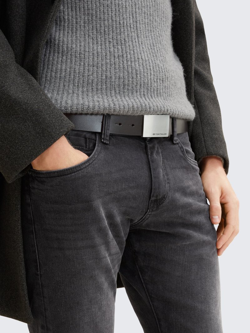 Pull gris côtelé, manteau noir et jean foncé, avec une ceinture noire élégante à boucle argentée métallique. Les accents incluent des textures subtiles.