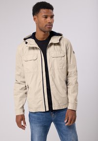 Béžová lehká bunda s kapucí, která má dvě náprsní kapsy a kontrastní navy podšívku. Materiál má hladkou strukturu a volný střih.