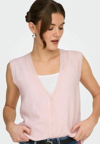 Femme portant un gilet sans manches rose pâle boutonné par-dessus un haut blanc, avec un jean bleu et des boucles d'oreilles argentées, regardant vers le bas et sur le côté.