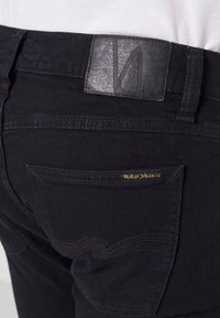 Černé džíny z denimu s texturovaným koženým pasem s raženým logem, zadní kapsa se zakřiveným prošíváním a zlatou vyšívanou značkovou etiketu.