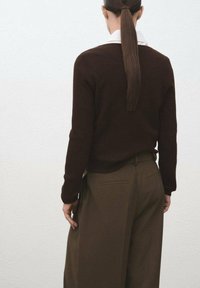 Persona con una larga coleta oscura, vistiendo un suéter marrón oscuro sobre una camisa blanca y pantalones anchos marrones, de pie contra una pared blanca.