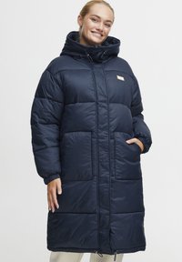 Navy blue puffer coat with a hood, featuring large quilted panels, side pockets, and adjustable drawstrings at the hem and neckline.