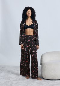 Ensemble de pyjama noir à imprimé floral avec un pantalon large et une chemise assortie, fabriqué en tissu doux. La chemise est déboutonnée sur un soutien-gorge en dentelle noire.