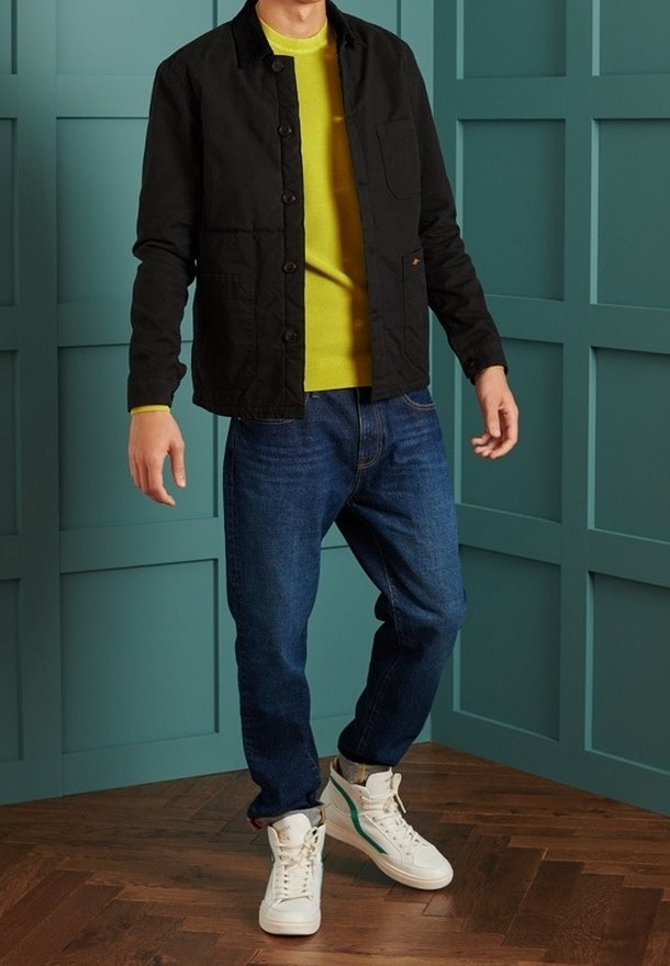 Veste noire boutonnée portée sur un pull jaune vif, associée à un jean bleu et des baskets blanches avec des accents verts. Design simple et coupe décontractée.