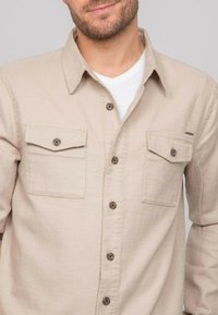 Chemise beige à boutons, fabriquée en tissu tissé, dotée de deux poches poitrine avec fermetures à bouton et d'un col doublé. Surface texturée.