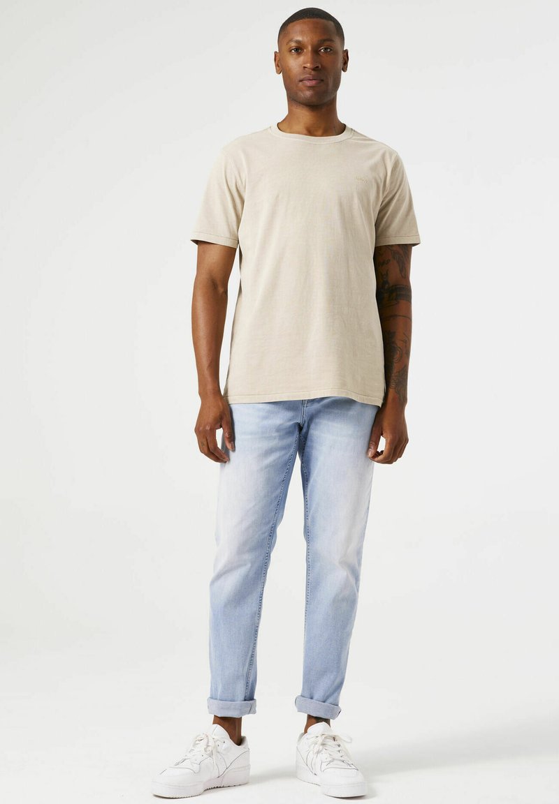 Camiseta de manga corta beige con cuello redondo, combinada con jeans de color azul claro y zapatillas blancas, creando un look casual.