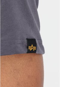 Primer plano de una camiseta de algodón gris que muestra una etiqueta negra con un emblema dorado en el dobladillo. Textura suave, diseño de manga corta, ajuste casual.