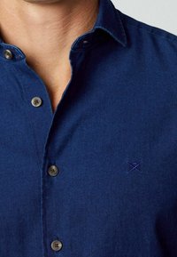 Primer plano de una persona que lleva una camisa de botones azul oscuro con un pequeño logo bordado en el pecho.
