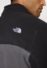 Sort og grå fleecejakke med høj krave. Har "The North Face" logoet broderet i hvidt på den øverste ryg. Tekstureret overflade.
