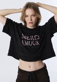 T-shirt nera cropped con tessuto texturizzato e arricciato e la scritta "MERCI AMOUR" stampata in rosa sulla parte anteriore. Maniche corte e ampie.