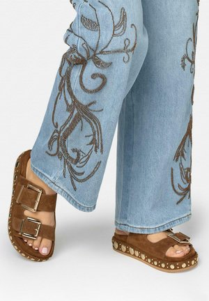 Sandales en daim marron avec double boucle et clous métalliques portées avec un jean bleu clair brodé de motifs floraux marron.