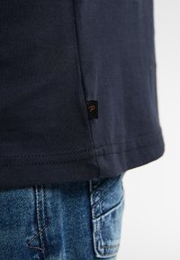 T-shirt in cotone blu navy con orlo curvo, caratterizzato da un piccolo etichetta nera con una "P" stilizzata sul lato vicino alla cucitura. Jeans in denim strappati visibili.