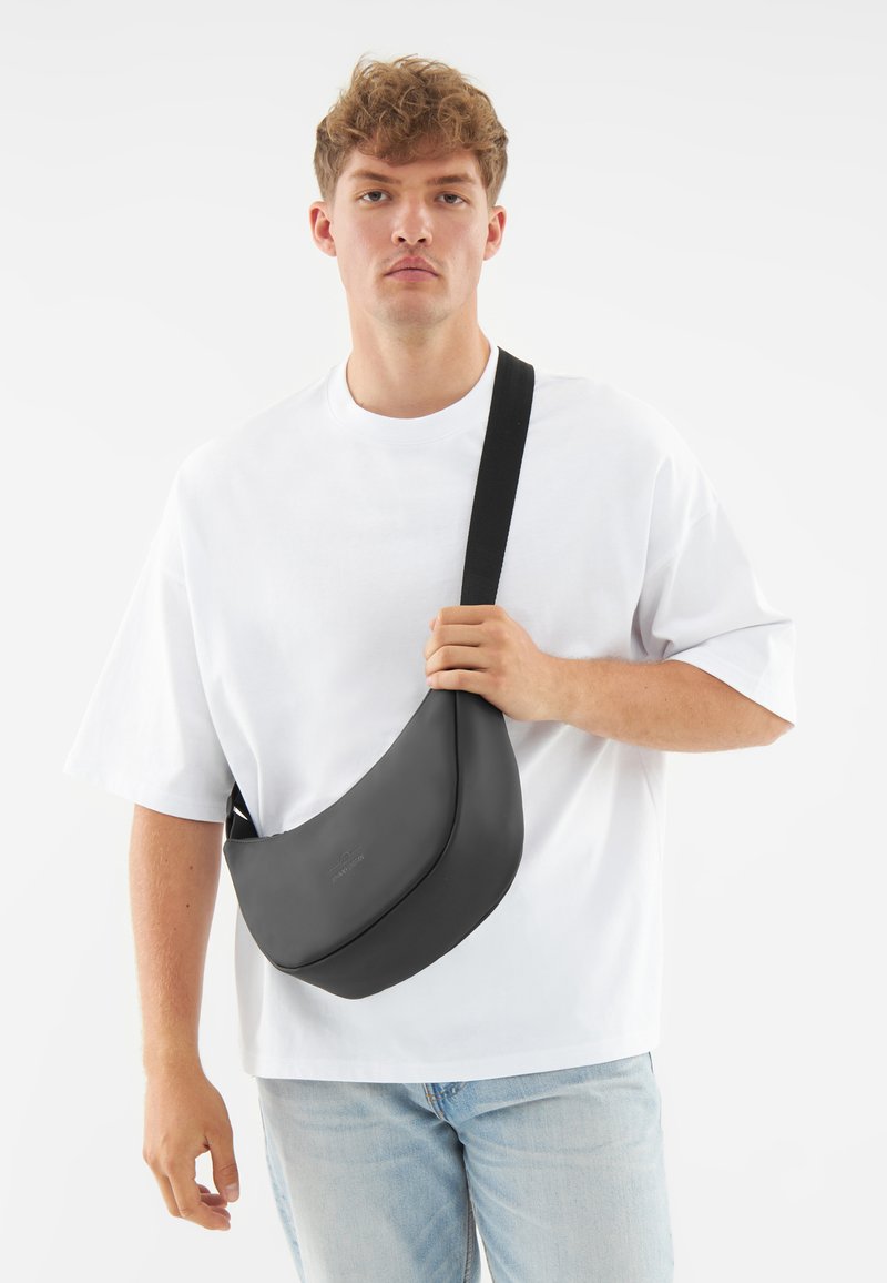 Borsa a spalla in pelle nera con un design liscio e curvo e tracolla nera regolabile, indossata sopra una t-shirt bianca e jeans azzurri chiari.