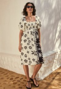 Mekko on neliönmuotoisella pääntiellä, lyhyillä puffihihnoilla ja sekoituksella mustia kukkakuosia vaaleassa taustassa; yhdistetty mustiin sandaaleihin.