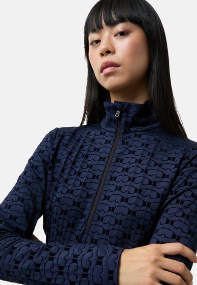 Námořnicky modrý pulovr na zip vyrobený z texturované látky se vzorem tvarovaných motivů. Stojací límec a přiléhavý střih s viditelným zipem.