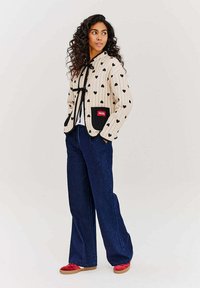 Béžová prešívaná bunda s čiernym vzorom srdca, čiernymi akcentmi, viazaným výstrihom a červenou visačkou; spojená s voľnými tmavomodrými džínsami a červenými topánkami.