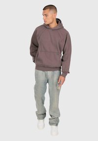 Młody mężczyzna patrzy w lewo, ubrany w fioletową bluzę z kapturem, jasne, przetarte dżinsy i białe sneakersy, stojąc na tle jasnego, gładkiego tła.