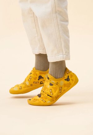 Gelbe Schuhe aus weichem Stoff, mit verspielten Tiermustern und einem Klettverschluss. Die Sohle ist strukturiert für besseren Halt, kombiniert mit gerippten, neutralen Socken.
