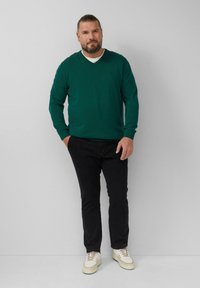 Zielony sweter w serek, w połączeniu z czarnymi spodniami i kremowymi sneakersami. Sweter ma gładką fakturę i luźny krój.