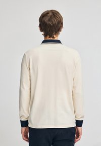 Hellcreme Langarm-Polohemd mit navyblauem Kragen und Bündchen. Weicher Stoff, minimalistisches Design und entspannter Schnitt, von hinten betrachtet.