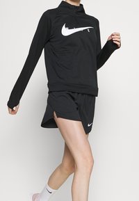 Musta Nike-urheilu pusero, jossa on puolivetoketju kauluksessa ja valkoinen logo, yhdistettynä matching mustiin shortsit, joissa on sivuaukot liikkumista varten.
