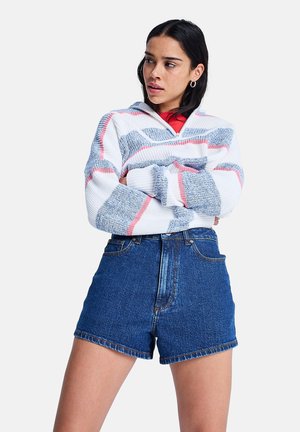Νεαρή γυναίκα φοράει λευκό, μπλε και ροζ ριγωτό πουλόβερ και κοντά, ψηλόμεσα μπλε τζιν σορτς, στέκεται με τα χέρια σταυρωμένα και κοιτάζει πλάγια.
