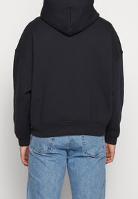 Personne portant un hoodie noir uni et un jean bleu clair, vue de dos sur un fond neutre.