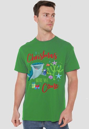 Mann trägt ein grünes T-Shirt mit bunten Unterwassercharakteren, Geschenken und dem Text "Weihnachten, wir kommen" in roter und grüner Schrift.