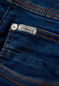 Sininen farkkukangas oranssilla ompelella, jossa on merkki "Lindbergh" ja nappiyksityiskohta. Sujuva rakenne, klassinen muotoilu.