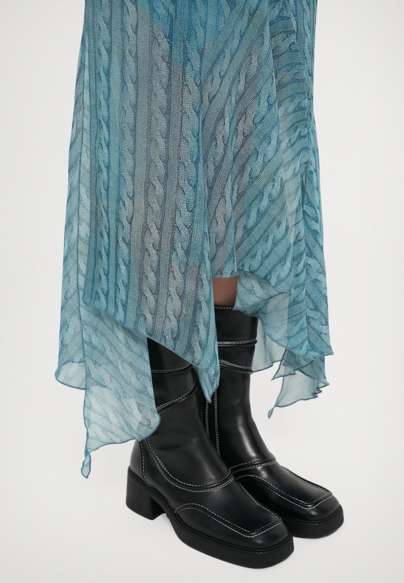 Black leather mid-calf boots with white stitching worn under a sheer teal dress with a wavy cable knit pattern and an asymmetrical hem.