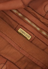 Interni in pelle marrone con una cerniera dorata e un'etichetta rettangolare che recita "CATERINA LUCCHI MADE IN ITALY." Finitura morbida e strutturata.