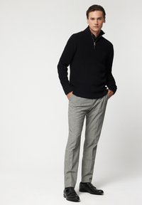 Suéter negro de punto con media cremallera, combinado con pantalones de cuadros grises claros y zapatos negros sin cordones. Detalles de corte sencillo y textura visibles.