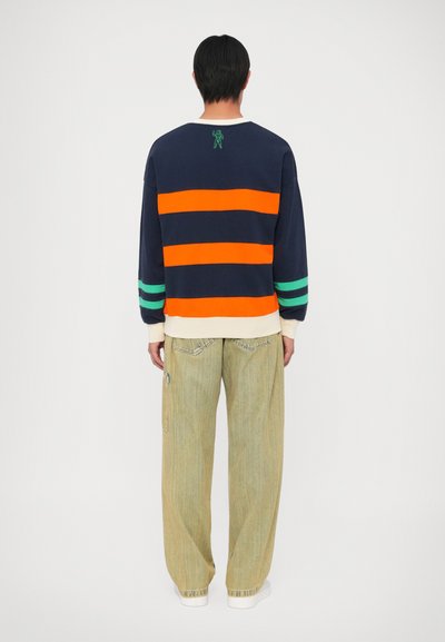 Sweatshirt azul-marinho com riscas laranja e verde, apresentando uma pequena figura verde bordada. Combinado com calças largas verde-claro.