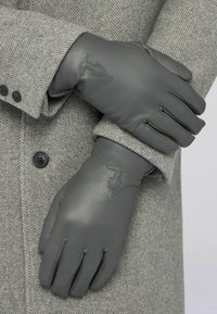 Gants en cuir gris présentés sur un manteau gris à motif chevrons. Ils comportent des accents cousus et un logo embossé au niveau du poignet.