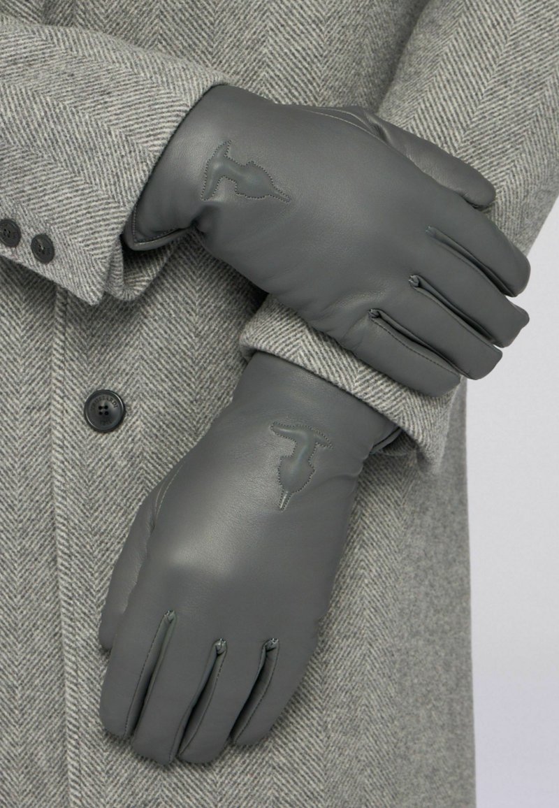 Gants en cuir gris présentés sur un manteau gris à motif chevrons. Ils comportent des accents cousus et un logo embossé au niveau du poignet.