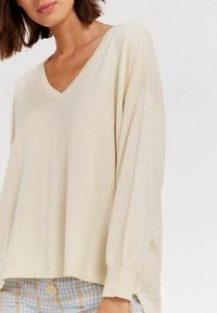 Pull à col en V crème, à coupe loose, avec des manches longues, des poignets côtelés et une texture douce, associé à un short à carreaux bleus.