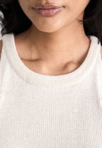 Απαλή μπεζ πλεκτή μπλούζα με στρογγυλή λαιμόκοψη και μικρές τιράντες, με υφή που δημιουργεί ένα ανάγλυφο σχέδιο πάνω στον ύφασμα.