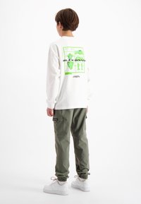 Langärmliges weißes Hemd mit grünen Grafikdrucks auf dem Rücken, kombiniert mit olivgrünen Cargo-Hosen und weißen Sneakers.