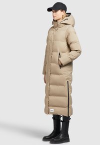 Kvinde iført en beige, lang pufferjakke med hætte, sort baseballkasket, sorte bukser og sorte støvler, står sidelæns på en grå baggrund.