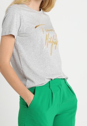 Nainen, jolla on yllään vaaleanharmaa Tommy Hilfiger -t-paita, jossa on kultakirjaus, ja kirkkaan vihreät korkeavyötäröiset housut, kädet rentoina taskuissa, valkoista taustaa vasten.